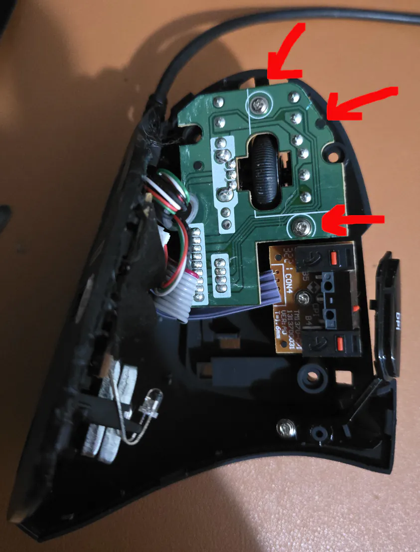 Inside of computer mouse. Three arrows are drawn to the screws and a block notch that hold the circuit board in place. You can see a mouse wheel in the center of the circuit board. 